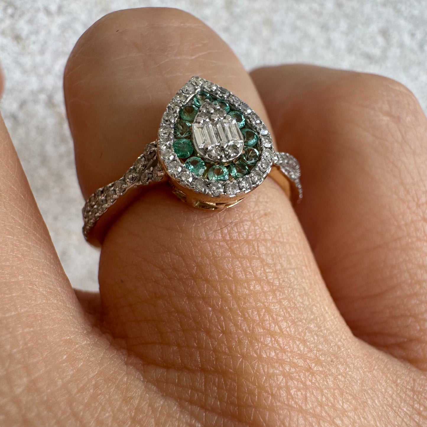 טבעת זהב צהוב 14 קראט בעיצוב טיפה (pear shape), משובצת יהלומים לבנים בשיבוץ הילה כפולה (double halo) ואבני אמרלד קטנות המקיפות מרכז ייחודי של יהלומים בחיתוך באגט