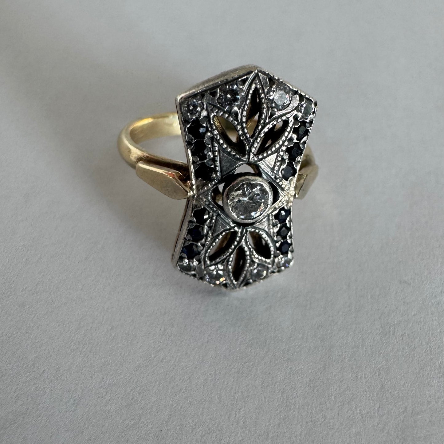 טבעת זהב 14 קראט בעיצוב אר-דקו גיאומטרי, משובצת יהלום מרכזי ומסגרת של אבני ספיר כהות ויהלומים נוצצים.