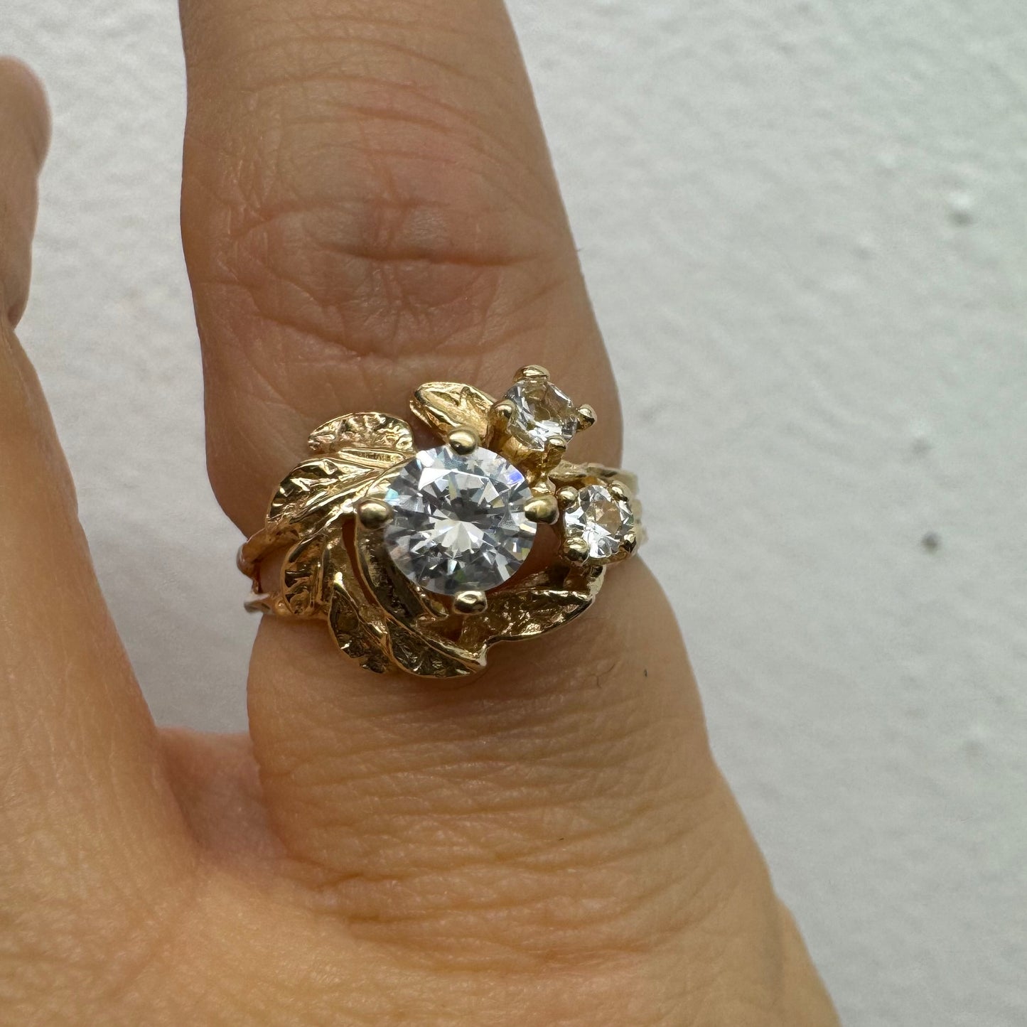 טבעת זהב צהוב 14 קראט בעלת עיצוב אורגני עשיר עם עלים מגולפים סביב שלוש אבנים שקופות נוצצות. (לא יהלומים)