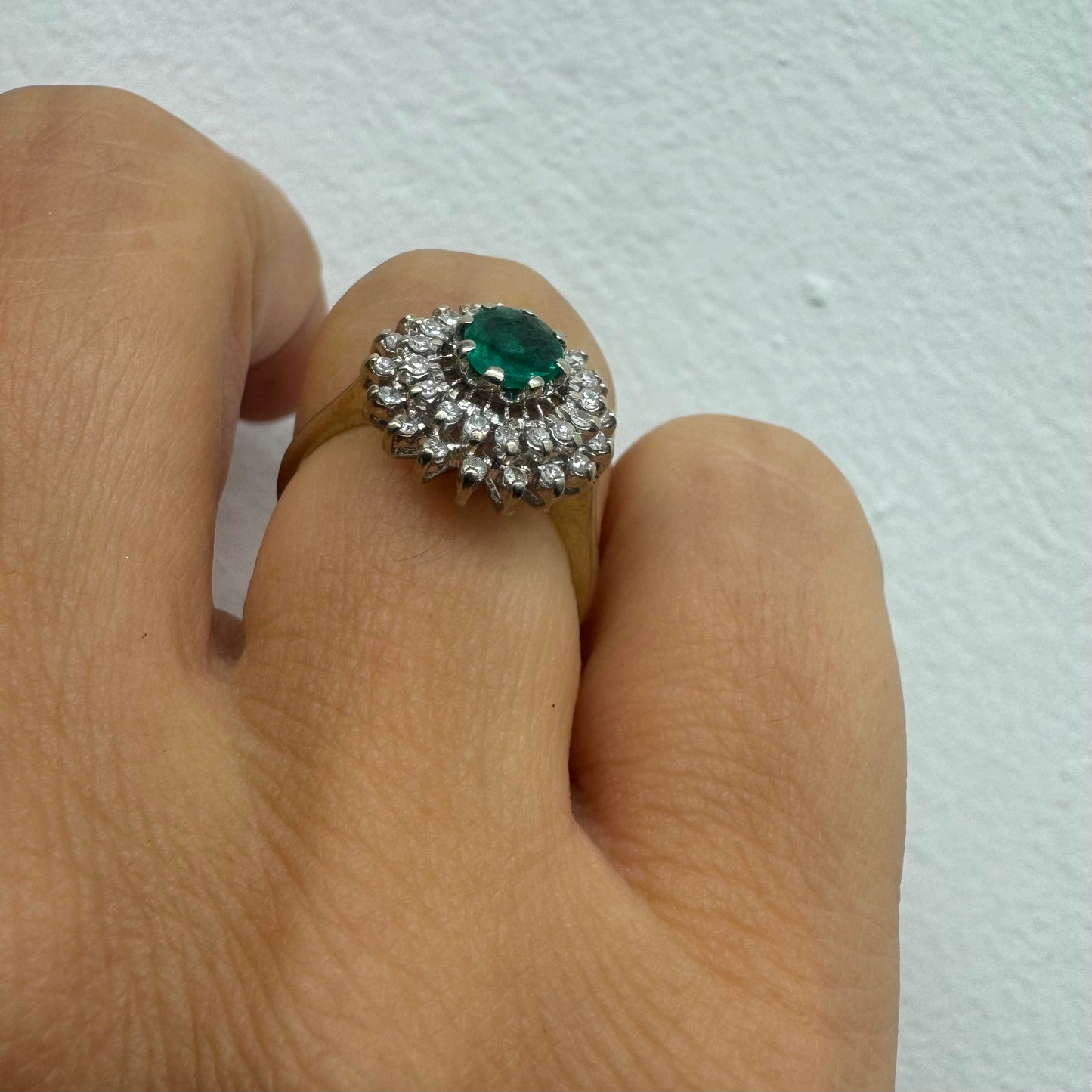 טבעת זהב לבן 14 קראט, משובצת אבן אמרלד ירוקה במרכז, מוקפת הילה עשירה של יהלומים בעיצוב קלאסי ואלגנטי. 