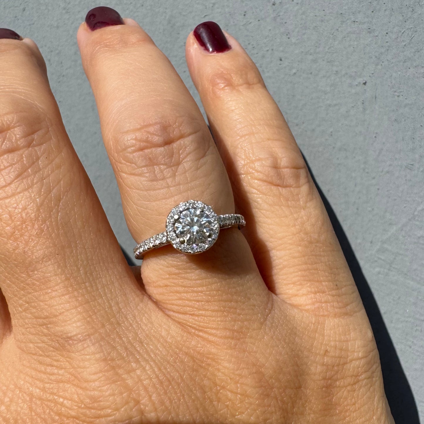 טבעת זהב לבן 14 קראט משובצת יהלום מרכזי עגול והילה נוצצת של יהלומים קטנים, עם יהלומים נוספים על החישוק.