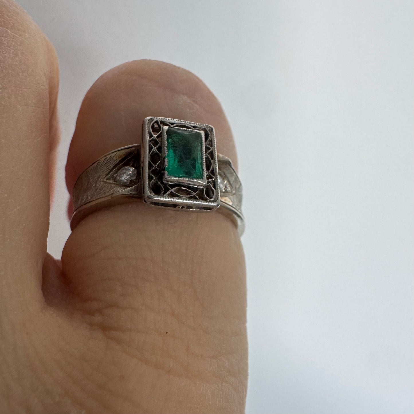 טבעת זהב לבן 14 קראט משובצת אבן אמרלד בחיתוך מלבני (Emerald cut) במרכז, מוקפת במסגרת מלבנית עם עבודת פיליגרן עדינה.