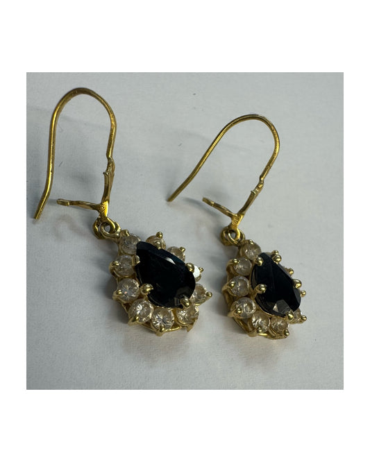 עגילי זהב צהוב 14 קראט בעיצוב קלאסי ומרשים, משובצים אבן ספיר בגוון כחול כהה בחיתוך טיפה ומסביבה אבנים שקופות עם הילה נוצצת שמוסיפה אור וזוהר לכל תנועה.