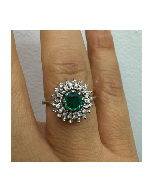 טבעת זהב לבן 14 קראט, משובצת אבן אמרלד ירוקה במרכז, מוקפת הילה עשירה של יהלומים בעיצוב קלאסי ואלגנטי. 